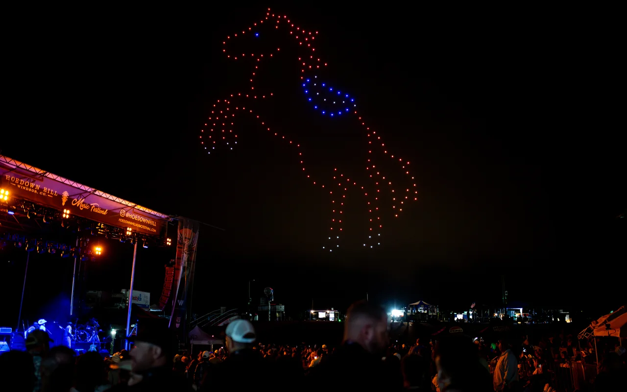A drone light show forms a rearing horse in red, white, and blue above a packed festival crowd at night.