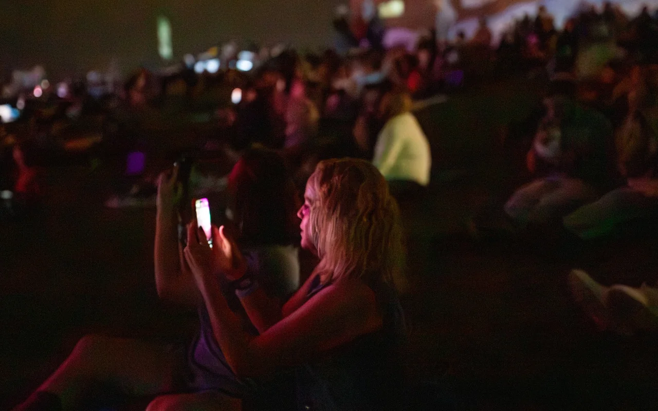 A woman films a drone light show on her phone amid a crowd seated on the grass, faces lit in the night.