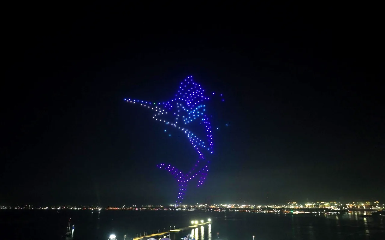 A drone light show forms a leaping marlin in blue above a shoreline of lights at night. Immersion produces drone light shows in Aspen, Colorado.