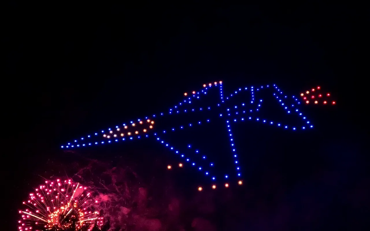 A drone light show formation glows in blue above bursting fireworks at night. Immersion produces drone light shows in Breckenridge, Colorado.