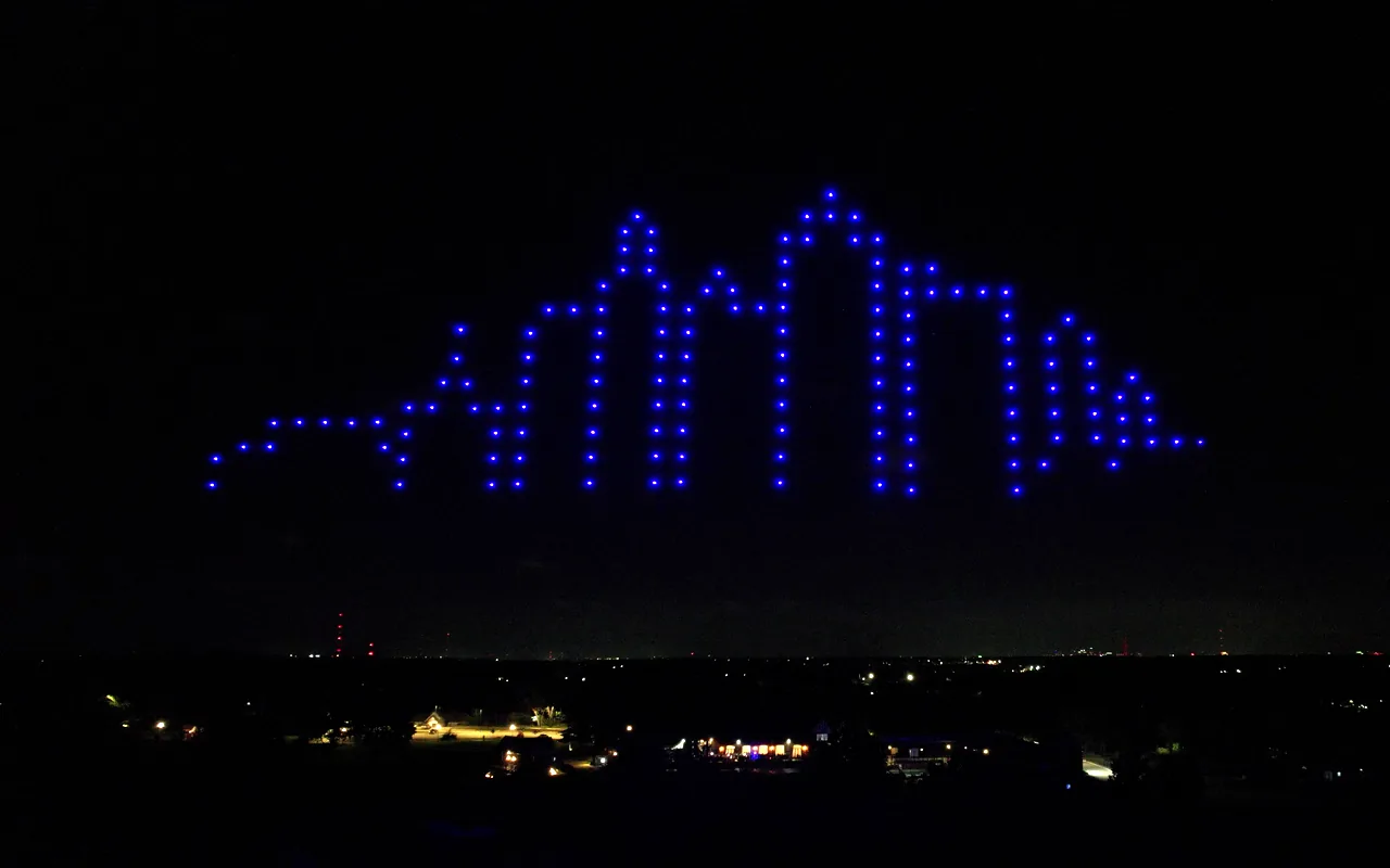 A drone light show forms a city skyline in blue above the lights of the metro at night. Immersion produces drone light shows in Kansas City, Missouri.