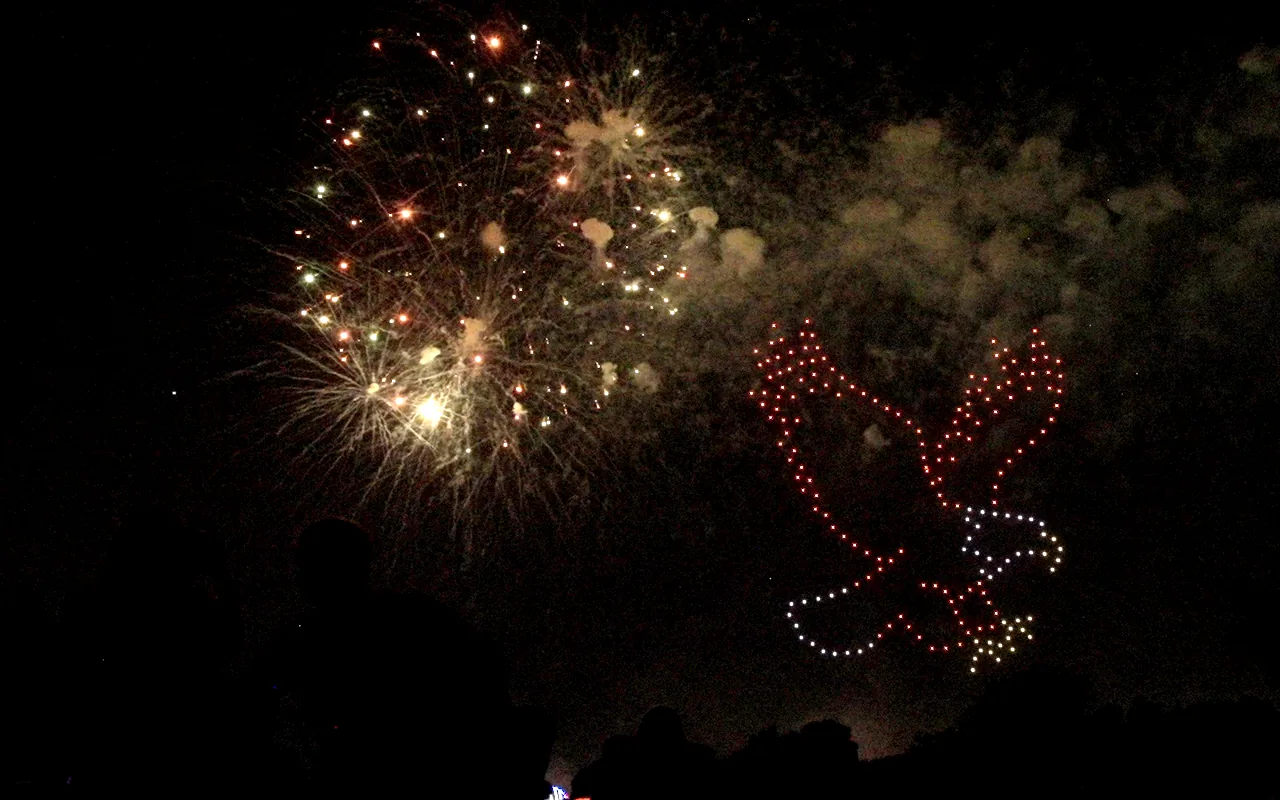 A drone light show forms an eagle beside a burst of fireworks at night. Immersion produces drone light shows in Loveland, Colorado.