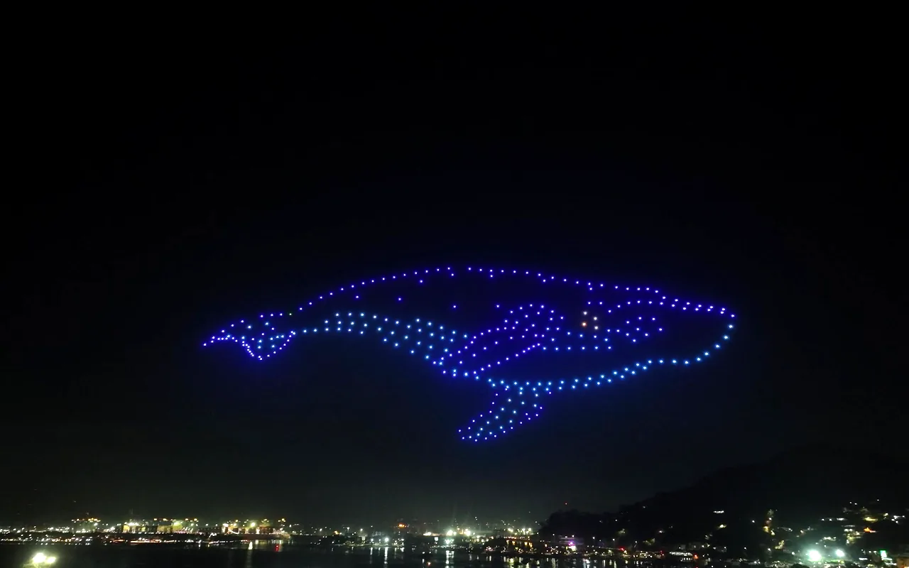 A drone light show forms a humpback whale in blue and white above a bay ringed with lights. Immersion produces drone light shows in Minneapolis, Minnesota.