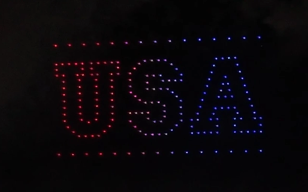 A drone light show spells USA in red, white, and blue above an Independence Day crowd.