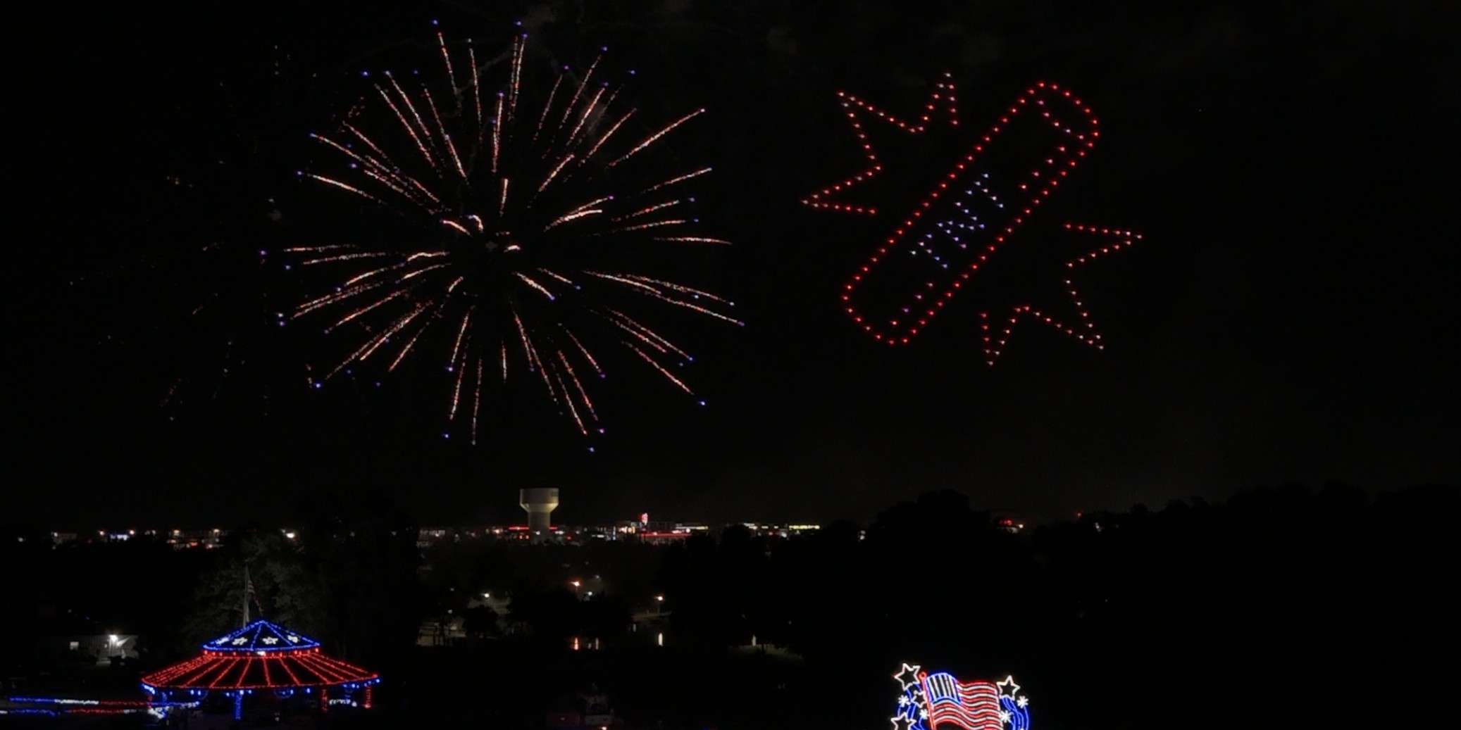 Fireworks burst beside a red, white, and blue drone formation over an Independence Day crowd, drones and fireworks in the same sky.