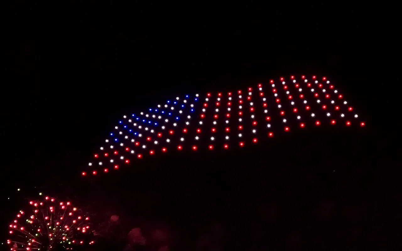 A drone light show forms the United States flag above a line of fireworks at night. Immersion produces drone light shows in St. Louis, Missouri.
