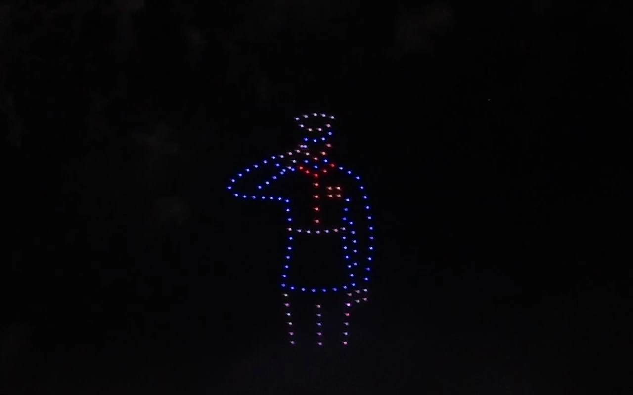 A drone light show forms a soldier saluting, lit in blue and red above the crowd at night.