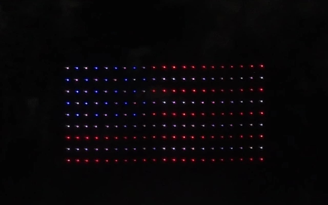 A drone light show forms a giant American flag in red, white, and blue over the crowd at night.