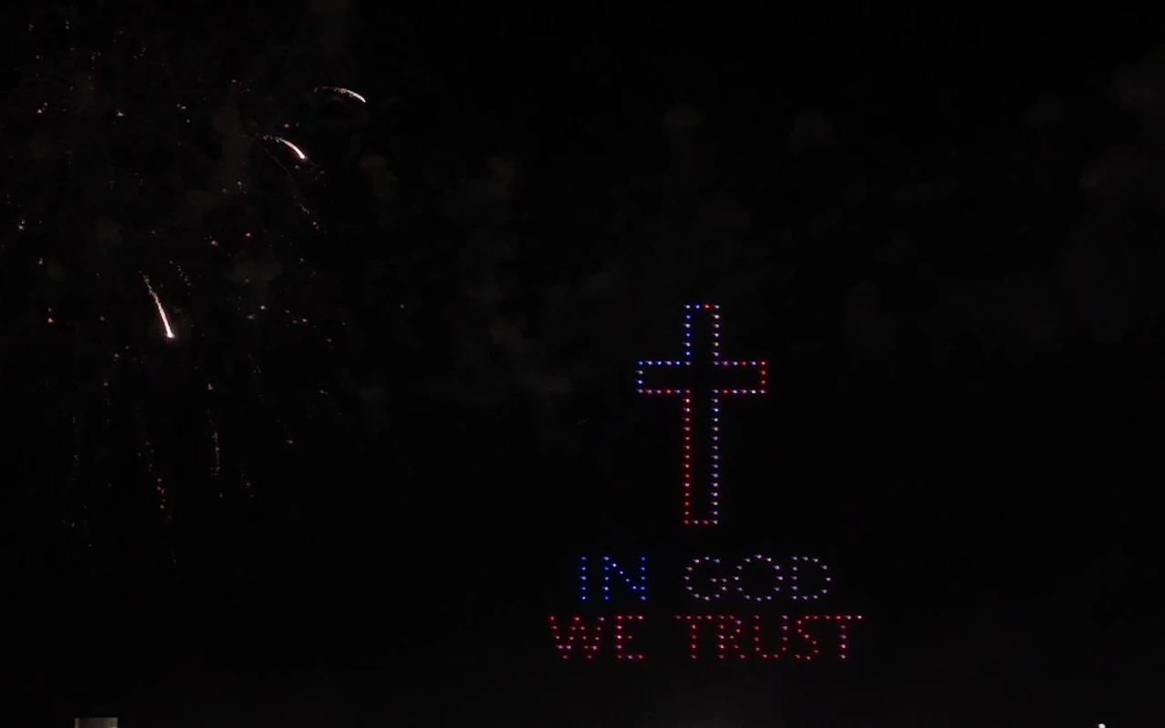 A drone light show forms a cross and the words In God We Trust in lights above the crowd at night.