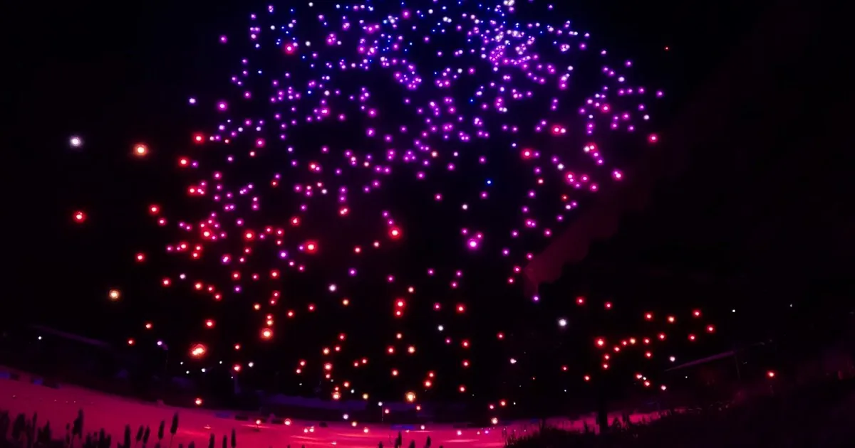 A drone light show seen from the ground at the back of the crowd, several hundred lights filling the night sky in magenta, violet and red above a venue lit pink below, with grass silhouetted in the foreground.
