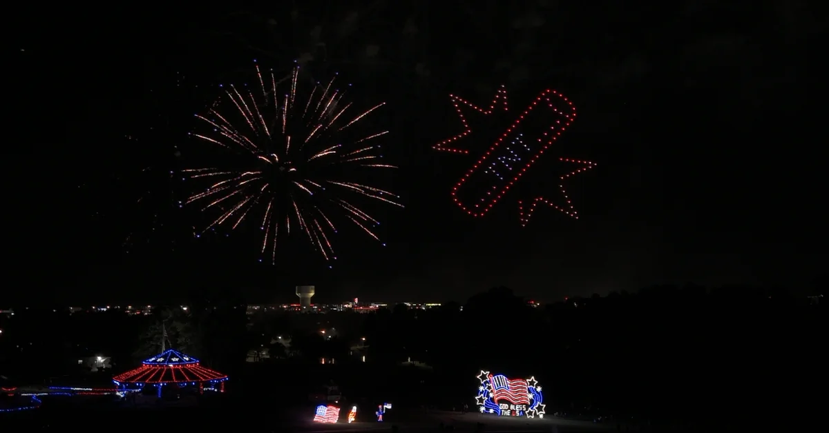 Fireworks bursting beside a red, white, and blue drone formation over an Independence Day crowd, drones and fireworks in the same sky.