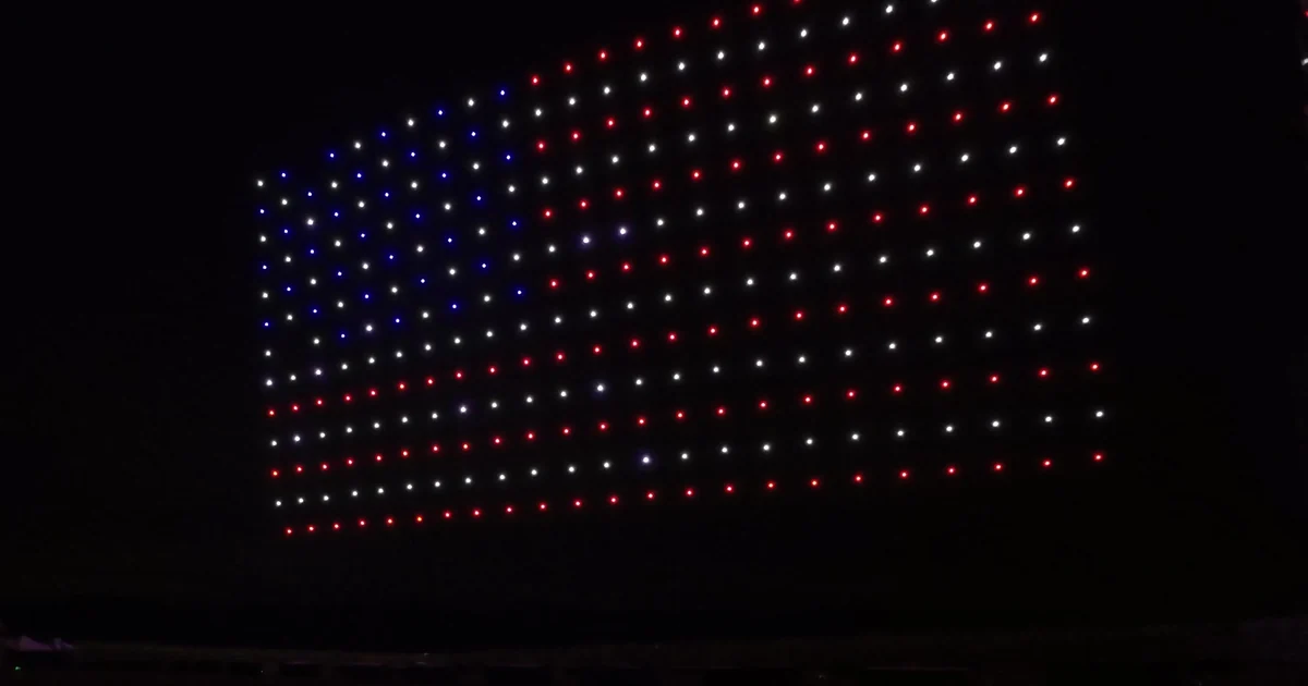 A drone light show forming the American flag against a black night sky, viewed from the ground at the audience's vantage point, with red, white, and blue lights held in a clean rectangular grid above a dark horizon.